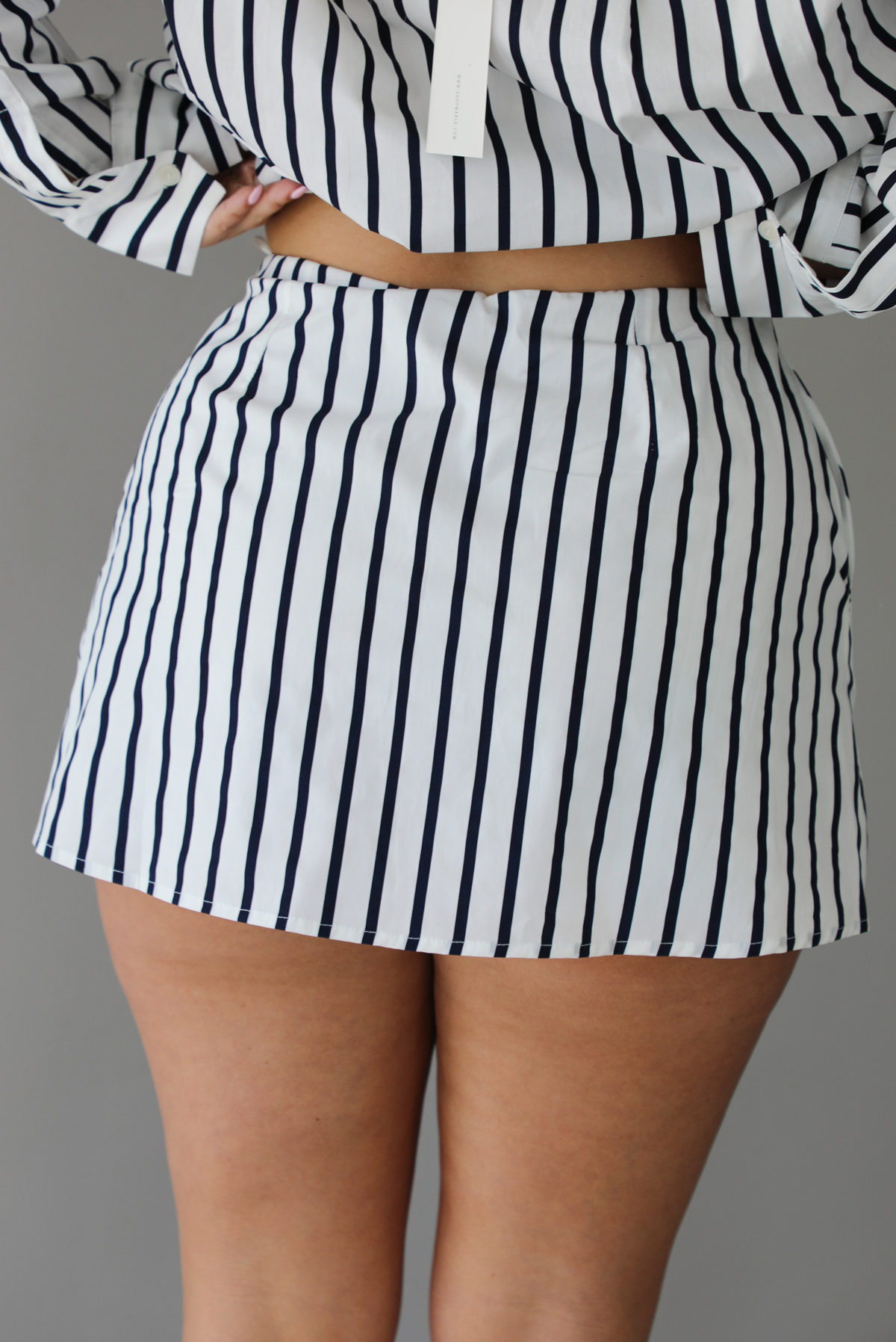 Woman wearing a white and navy striped set with a collared button-down top and mini skirt indoors against a white wall and shows a closeup of the back of the skirt. 