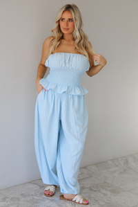 Woman wearing a blue two piece set featuring a strapless smocked top with a ruffled hem and high waisted flared pants with functional pockets on either side. The photo is taken indoors against a white wall with white marble flooring. 