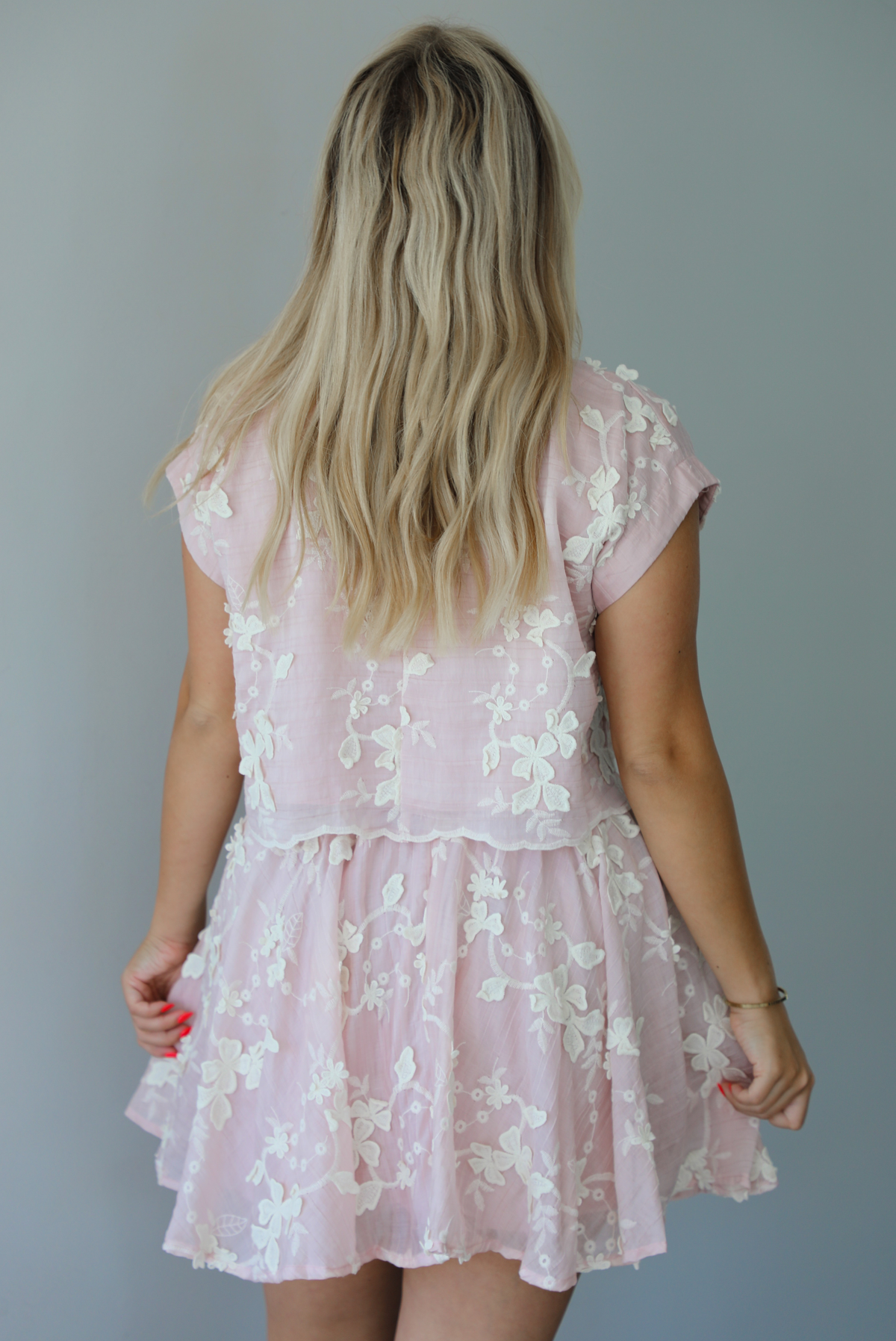 Woman wearing a pink and white floral embroidered two piece set featuring a cropped loose fit top and high waisted skirt indoors against a white wall. The photo shows the back of the set. 