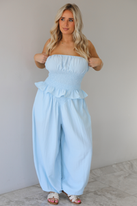 Woman wearing a blue two piece set featuring a strapless smocked top with a ruffled hem and high waisted flared pants with functional pockets on either side. The photo is taken indoors against a white wall with white marble flooring. 