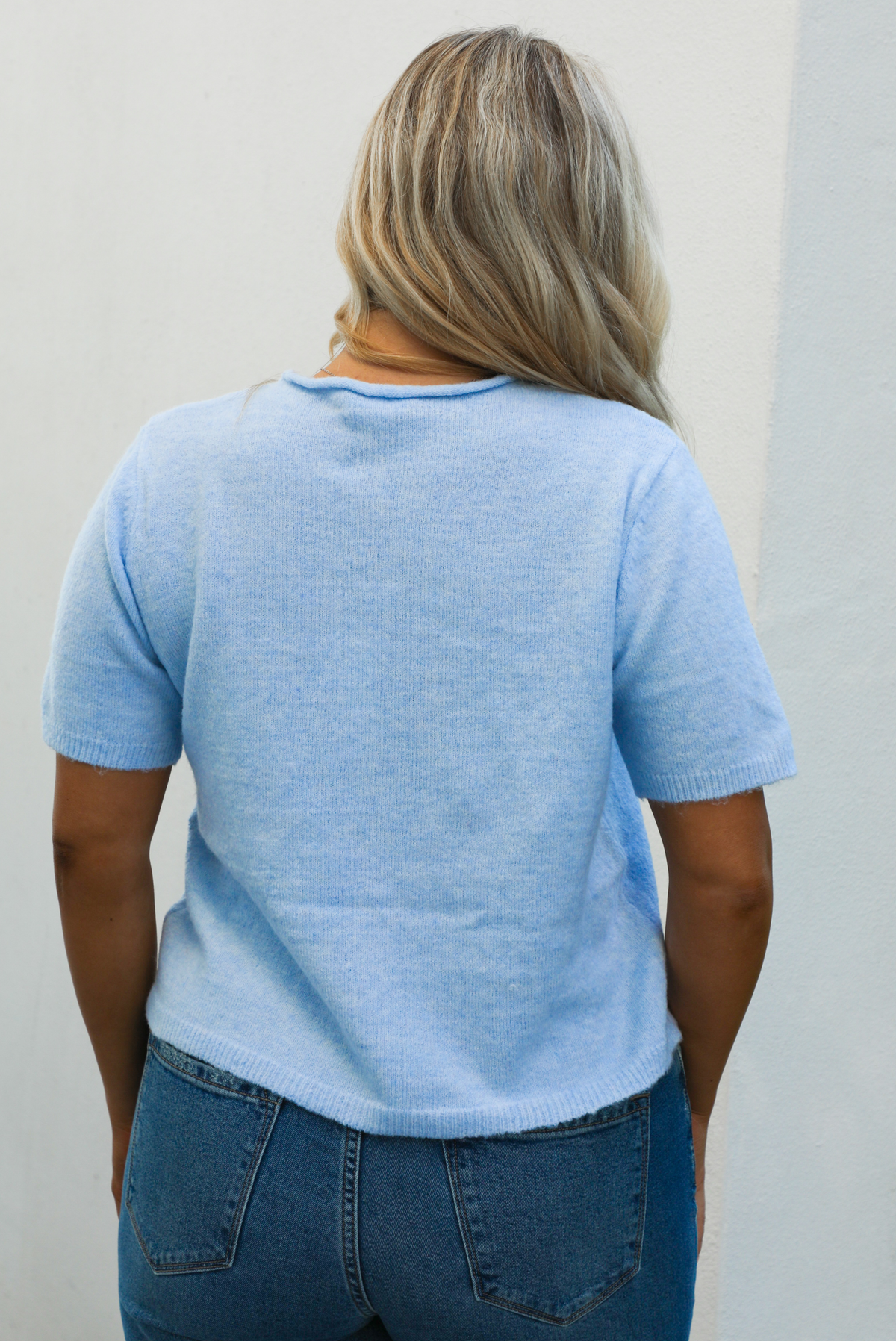 Back of a woman model wearing a baby blue knit top that has short sleeves, a relaxed fit, and functional button closures in the front paired with a medium wash denim. The photo is taken outdoors against a white wall with some greenery in the back. 