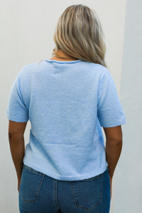 Back of a woman model wearing a baby blue knit top that has short sleeves, a relaxed fit, and functional button closures in the front paired with a medium wash denim. The photo is taken outdoors against a white wall with some greenery in the back. 
