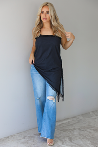 Woman wearing a charcoal asymmetrical hem top with a white polka dot print pattern and black lace detailing and medium wash denim jeans indoors against  a white wall with white marble flooring. 