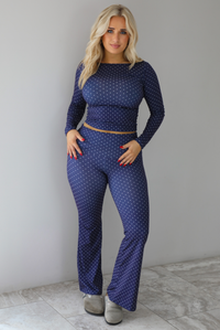 Woman wearing a navy two piece set that has a polka dot print pattern throughout.. The two piece set features high neckline, long sleeves, is fitted, and has a cropped length. The pants have a high waisted design with a fold over detail at the waistline and flared bottoms. The photo is taken indoors against a white wall with white marble flooring.