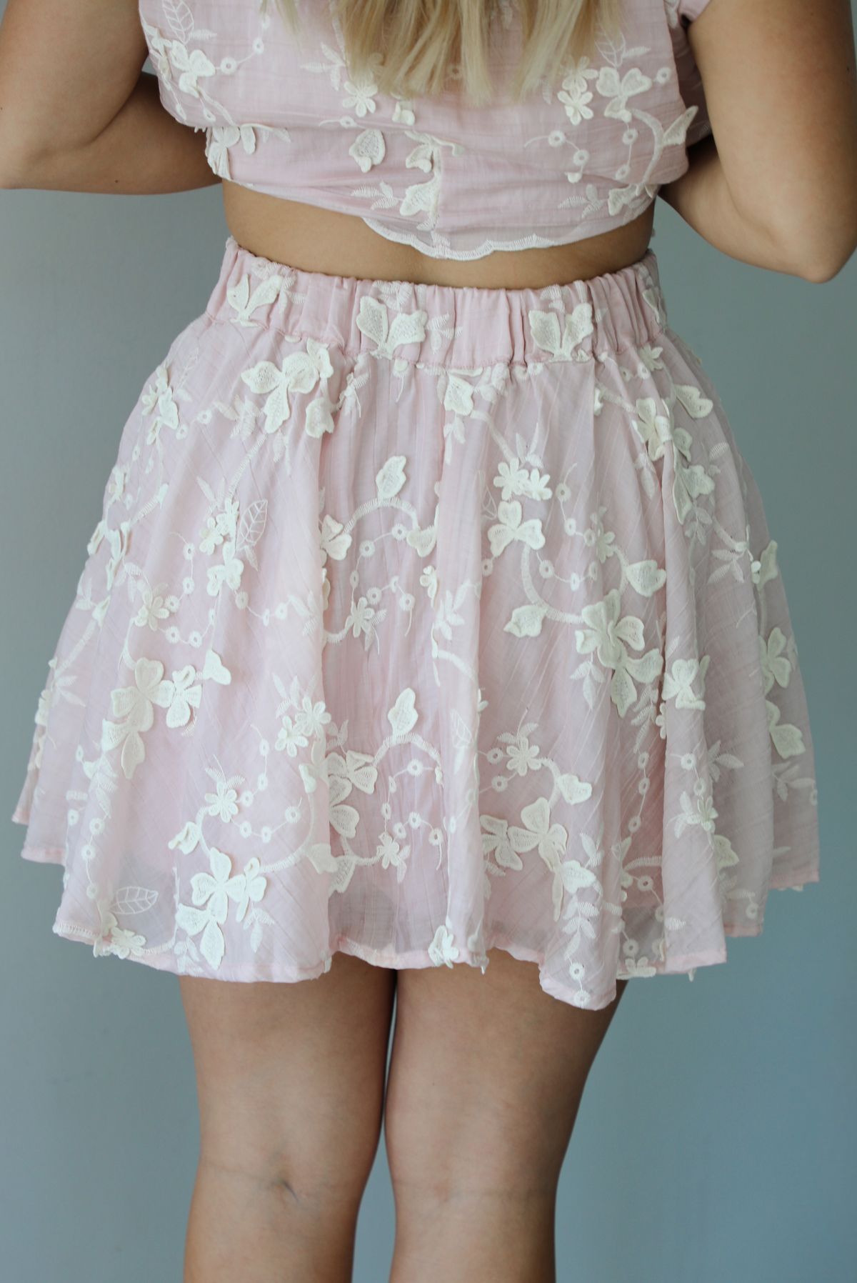 Woman wearing a pink and white floral embroidered two piece set featuring a cropped loose fit top and high waisted skirt indoors against a white wall. The photo shows a closeup of the back of the skirt, which has an elastic waistline.
