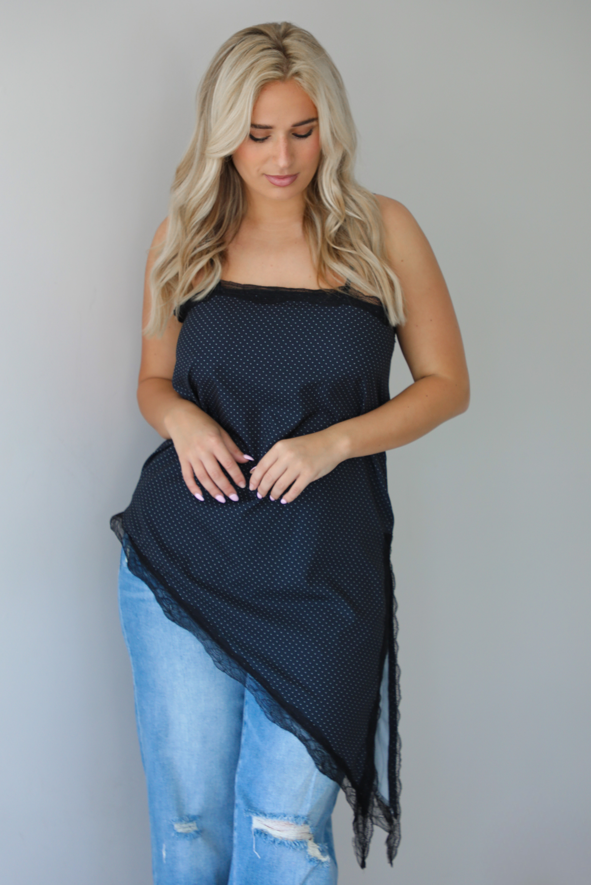 Woman wearing a charcoal asymmetrical hem top with a white polka dot print pattern and black lace detailing and medium wash denim jeans indoors against  a white wall.