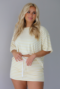 Woman wearing a yellow and white striped two piece set featuring an oversized two and fitted mini skirt indoors against a white wall.