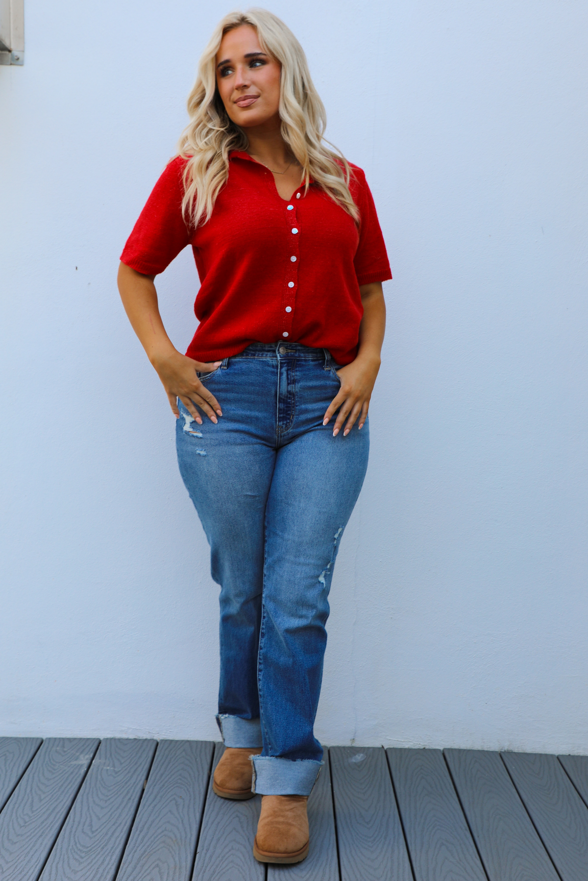 Woman model wearing a red knit top that has short sleeves, a relaxed fit, and functional button closures in the front paired with a medium wash denim. The photo is taken outdoors against a white wall.