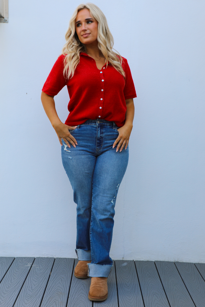 Woman model wearing a red knit top that has short sleeves, a relaxed fit, and functional button closures in the front paired with a medium wash denim. The photo is taken outdoors against a white wall.