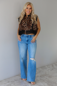Woman wearing a black and brown leopard print flyway top featuring eyelet lace detailing at the top of the neckline, sleeves, and hem with functional button closures with a mid-rise, medium wash denim jeans indoors against a white wall with white marble flooring. 