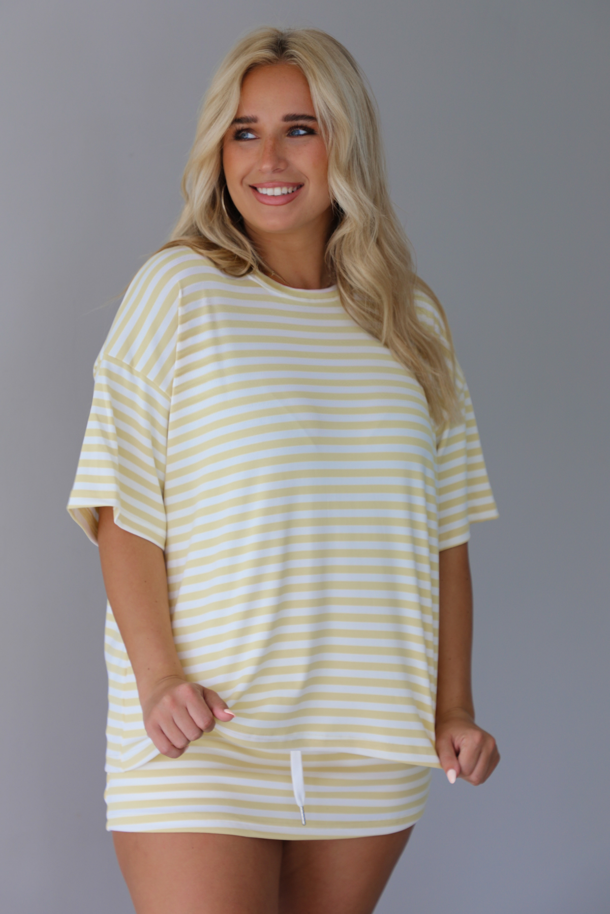 Woman wearing a yellow and white striped two piece set featuring an oversized two and fitted mini skirt indoors against a white wall. 