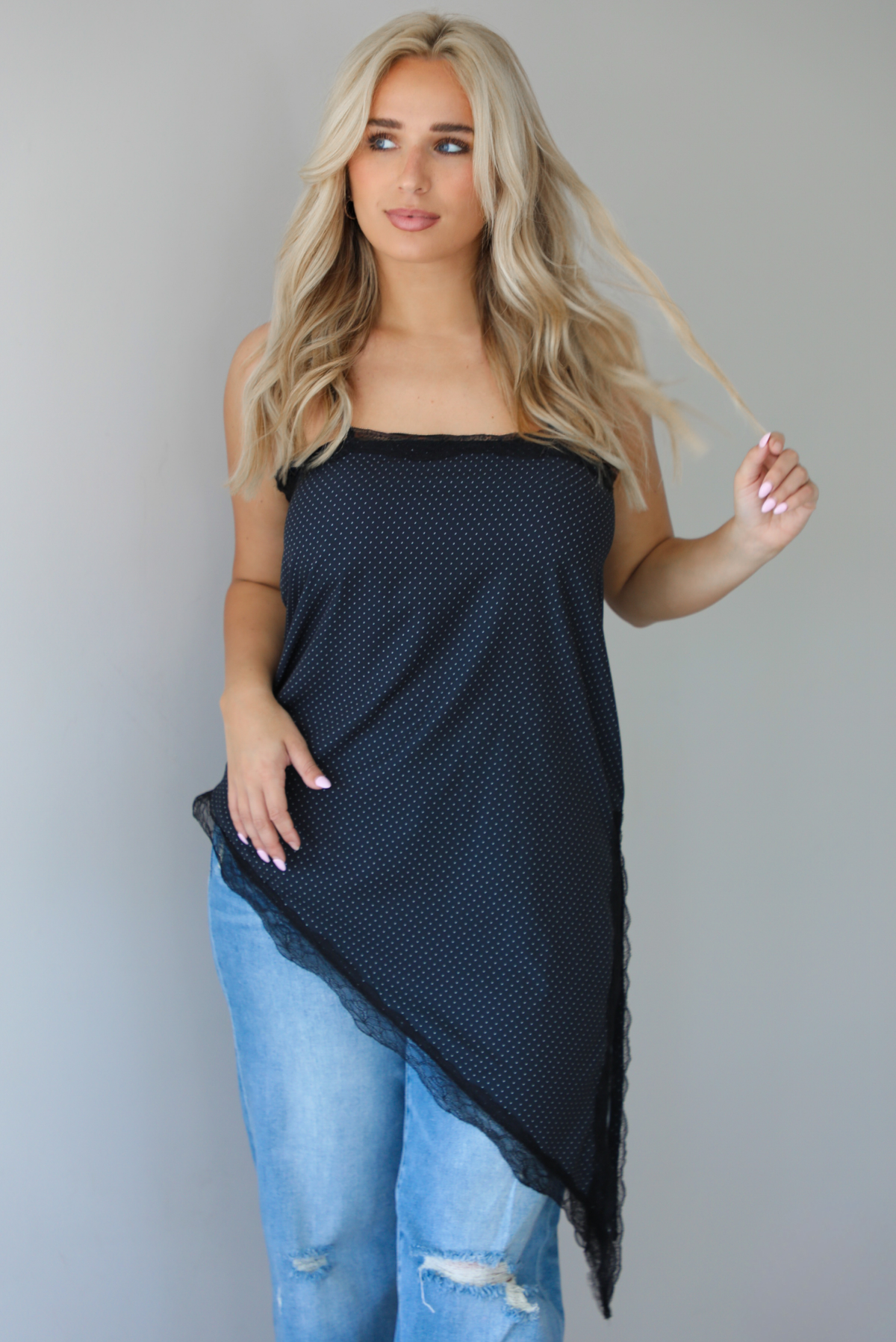 Woman wearing a charcoal asymmetrical hem top with a white polka dot print pattern and black lace detailing and medium wash denim jeans indoors against  a white wall.