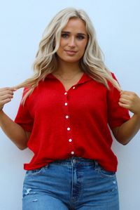 Close up of a woman model wearing a red knit top that has short sleeves, a relaxed fit, and functional button closures in the front paired with a medium wash denim. The photo is taken outdoors against a white wall.