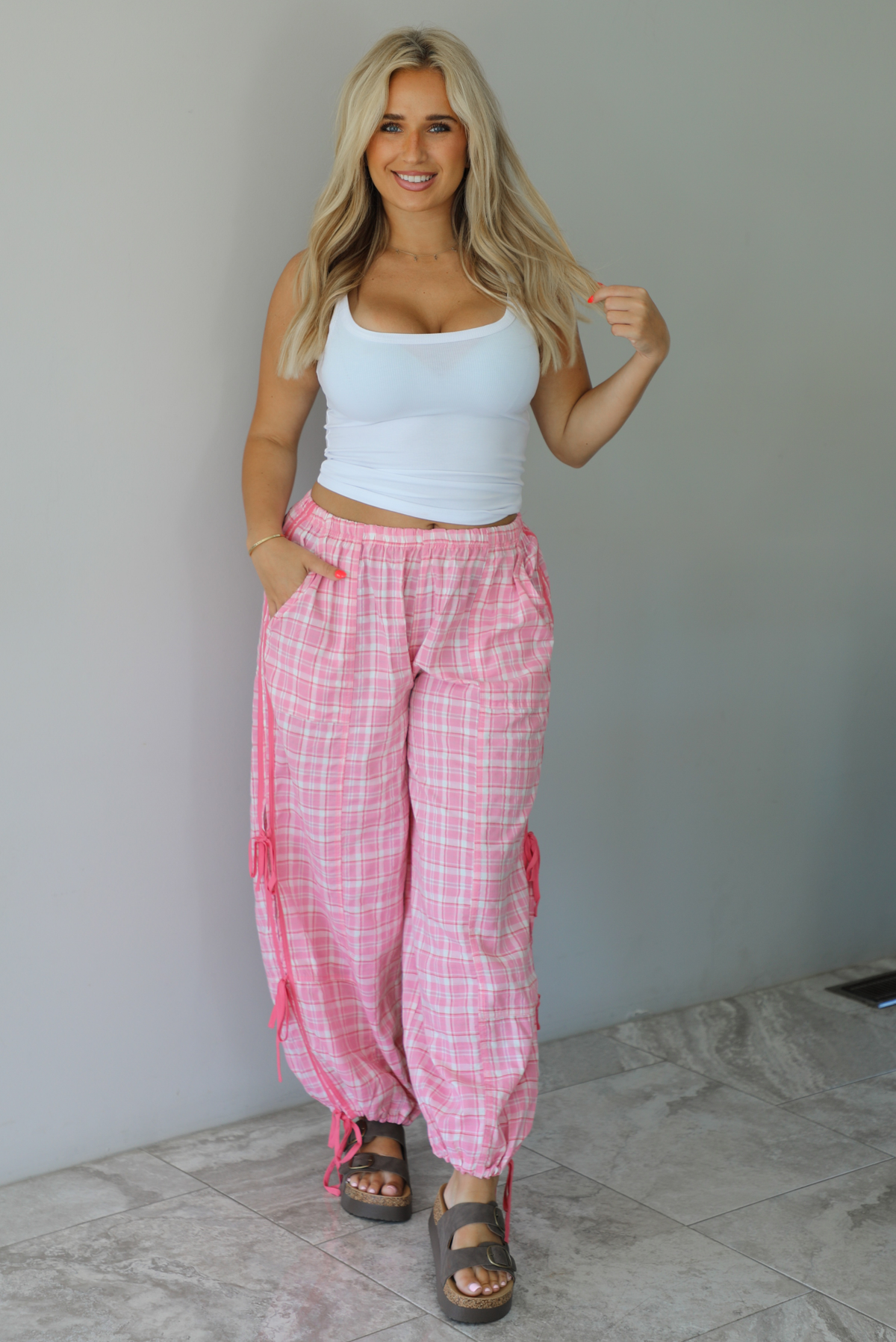 Woman wearing a white tank top with pink and white plaid pants that have a high, elastic waistline, functional pockets, and tie details on either side. The photo is taken indoors against a white wall with white marble flooring.