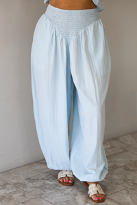 Woman wearing a blue two piece set featuring a strapless smocked top with a ruffled hem and high waisted flared pants with functional pockets on either side. The photo is taken indoors against a white wall and shows the front of the pants.
