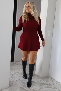 Woman model wearing a maroon colored sweater dress that has a high neckline, long sleeves, a ribbed texture, and stops on the upper thigh on the 5'7 model. The photo is taken indoors in an archway.