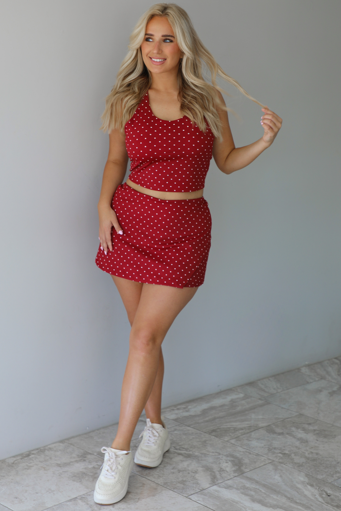 Woman wearing a red athleisure set with a white a white polka dot pattern throughout with a cropped tank and mini length skirt indoors against a white wall with white marble flooring. 