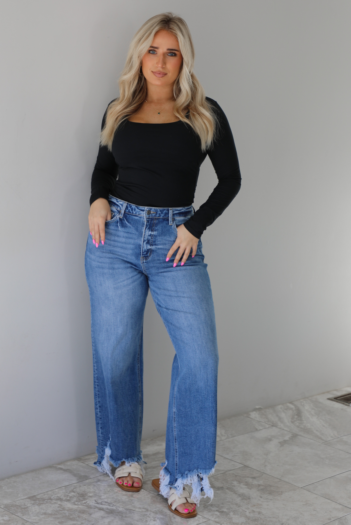 Woman wearing a black long sleeved fitted top tucked into high waisted distressed denim. The photo is taken indoors against a white wall with white marble flooring. 