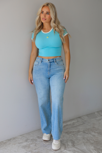 Woman wearing a blue crop top with an embroidered lemon on the front with mid-rise light-wash denim jeans against a white wall with white marble flooring. 