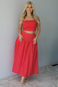 Woman wearing a red with white polka dots set featuring a cropped tank and high waisted maxi skirt indoors against a white wall with white marble flooring. 