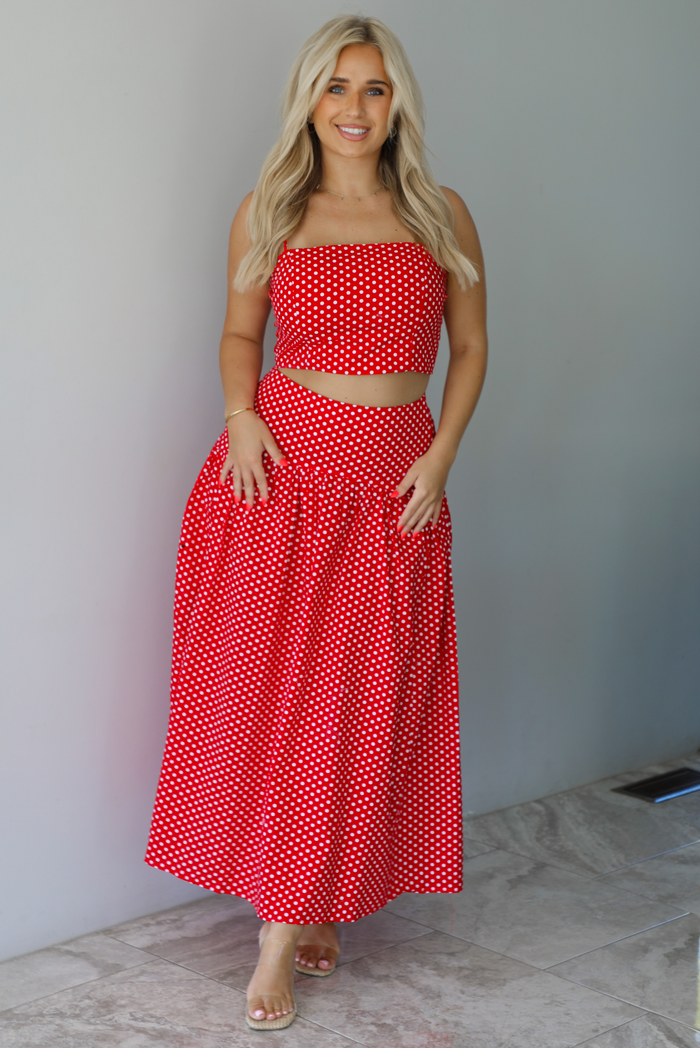 Woman wearing a red with white polka dots set featuring a cropped tank and high waisted maxi skirt indoors against a white wall with white marble flooring. 