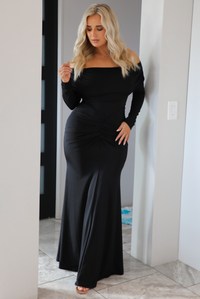 Woman wearing a fitted, black maxi dress that has a fold over detail at the shoulders, long sleeves, and a mermaid style skirt that's fitted at the hips and flows at the hem. The photo is worn indoors in a modern white home in an archway.