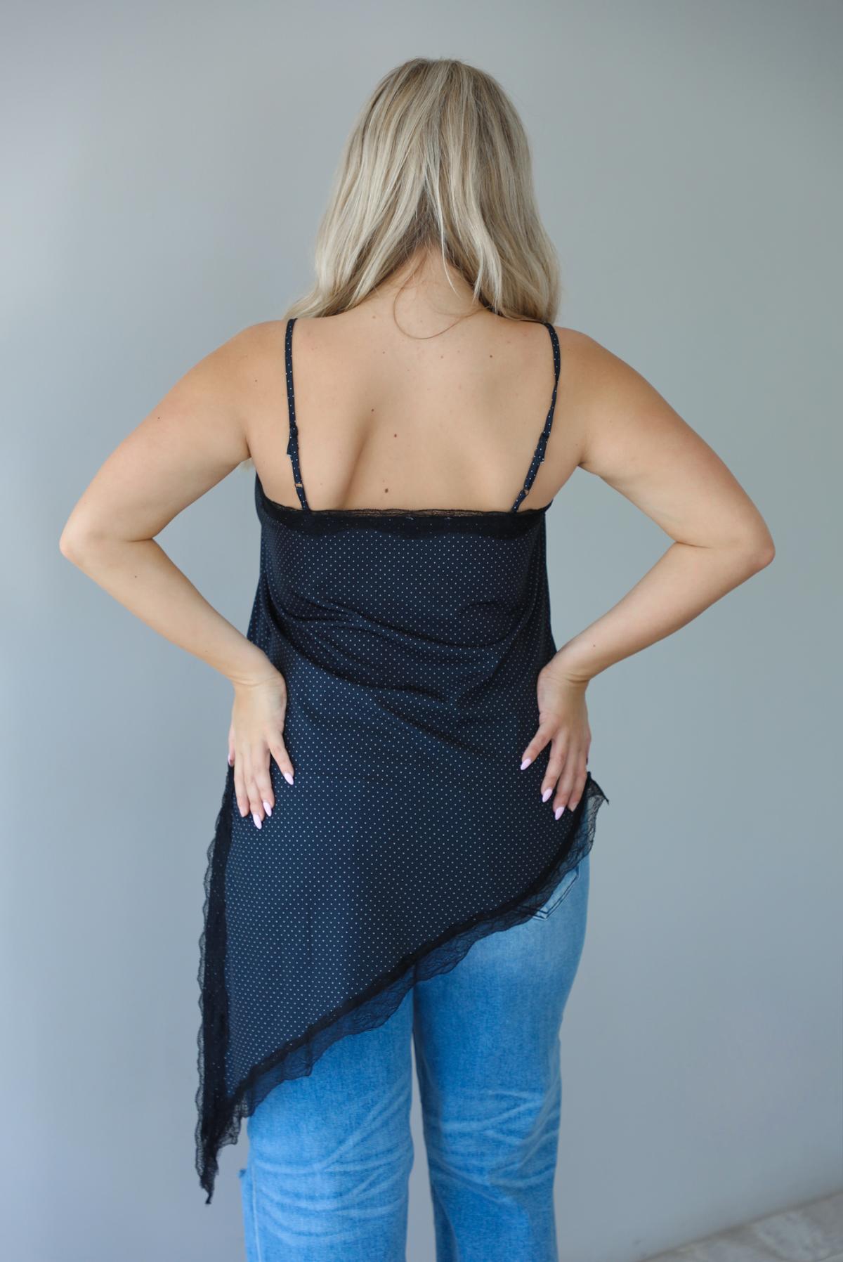 Woman wearing a charcoal asymmetrical hem top with a white polka dot print pattern and black lace detailing and medium wash denim jeans indoors against  a white wall and shows the back of the top. 