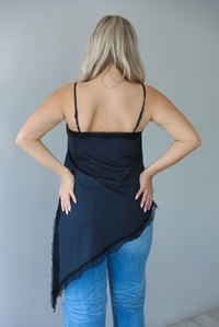 Woman wearing a charcoal asymmetrical hem top with a white polka dot print pattern and black lace detailing and medium wash denim jeans indoors against  a white wall and shows the back of the top. 