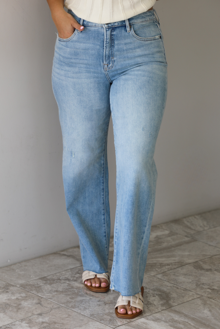 Woman wearing a pair of mid-rise light-wash denim jeans that have a raw, unfinished hem in front of a white wall with white marble flooring. 