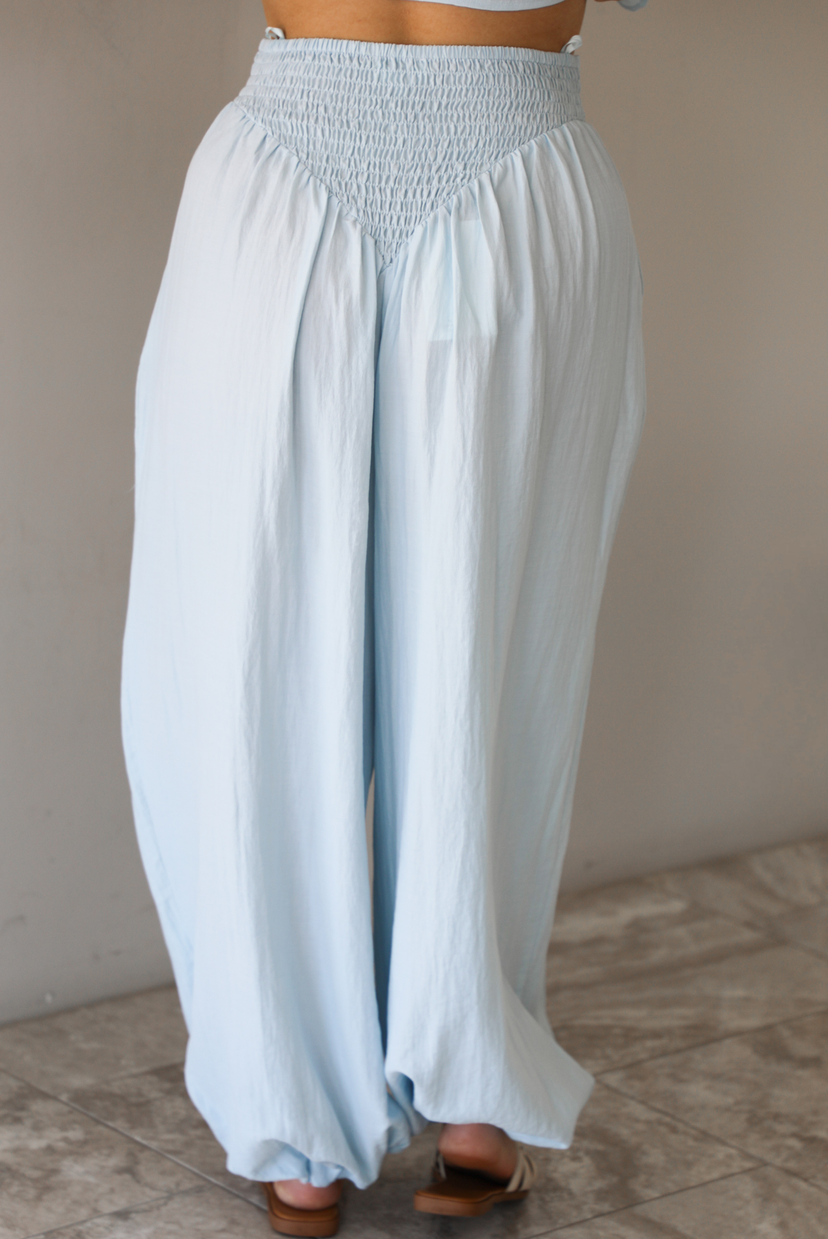 Woman wearing a blue two piece set featuring a strapless smocked top with a ruffled hem and high waisted flared pants with functional pockets on either side. The photo is taken indoors against a white wall and shows the back of the pants.
