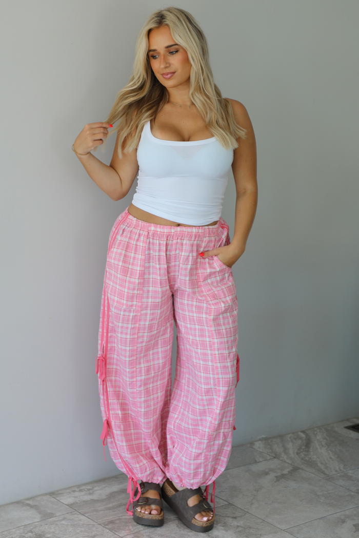 Woman wearing a white tank top with pink and white plaid pants that have a high, elastic waistline, functional pockets, and tie details on either side. The photo is taken indoors against a white wall with white marble flooring.