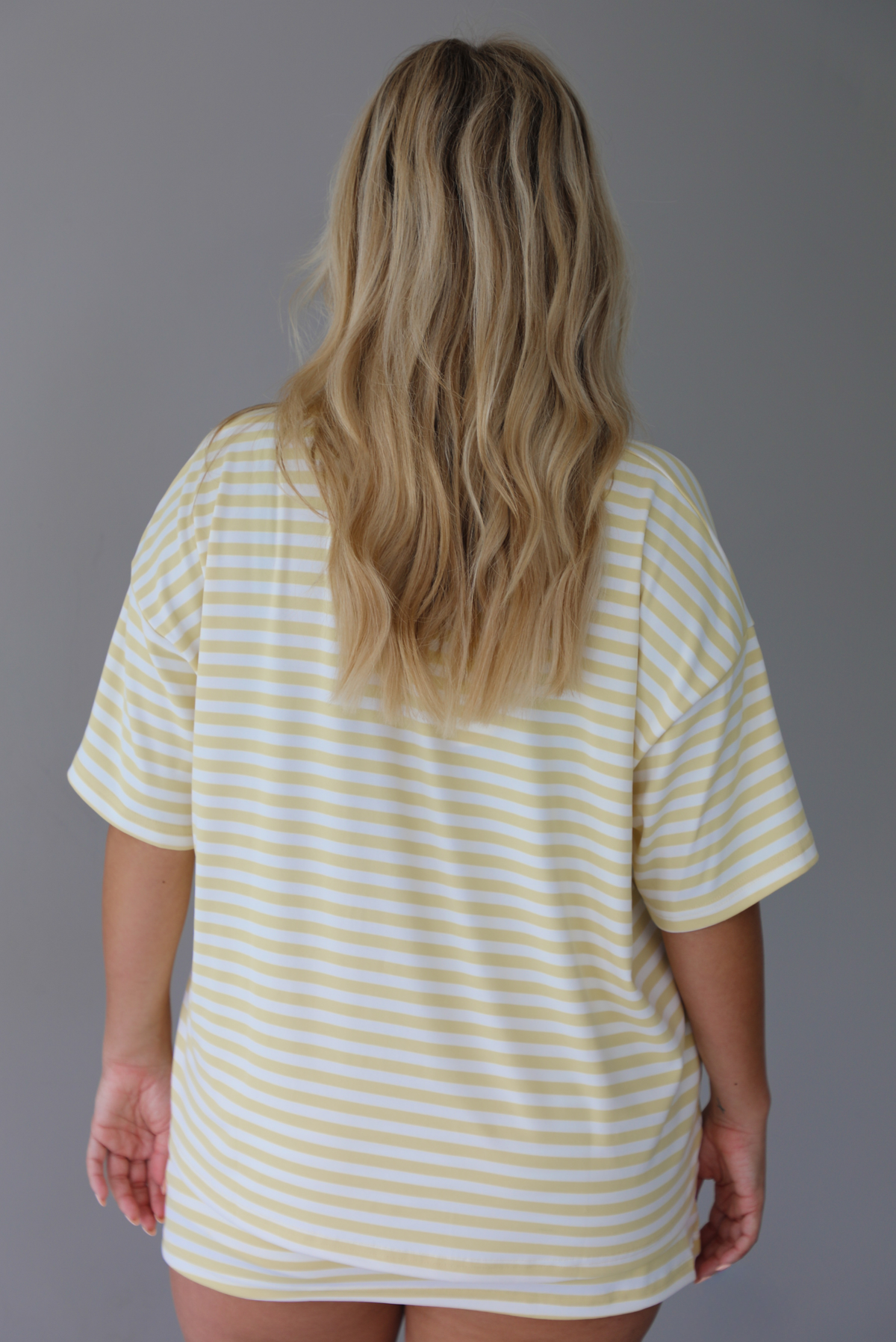 Woman wearing a yellow and white striped two piece set featuring an oversized two and fitted mini skirt indoors against a white wall. The photo shows a closeup of the back of the tee. 
