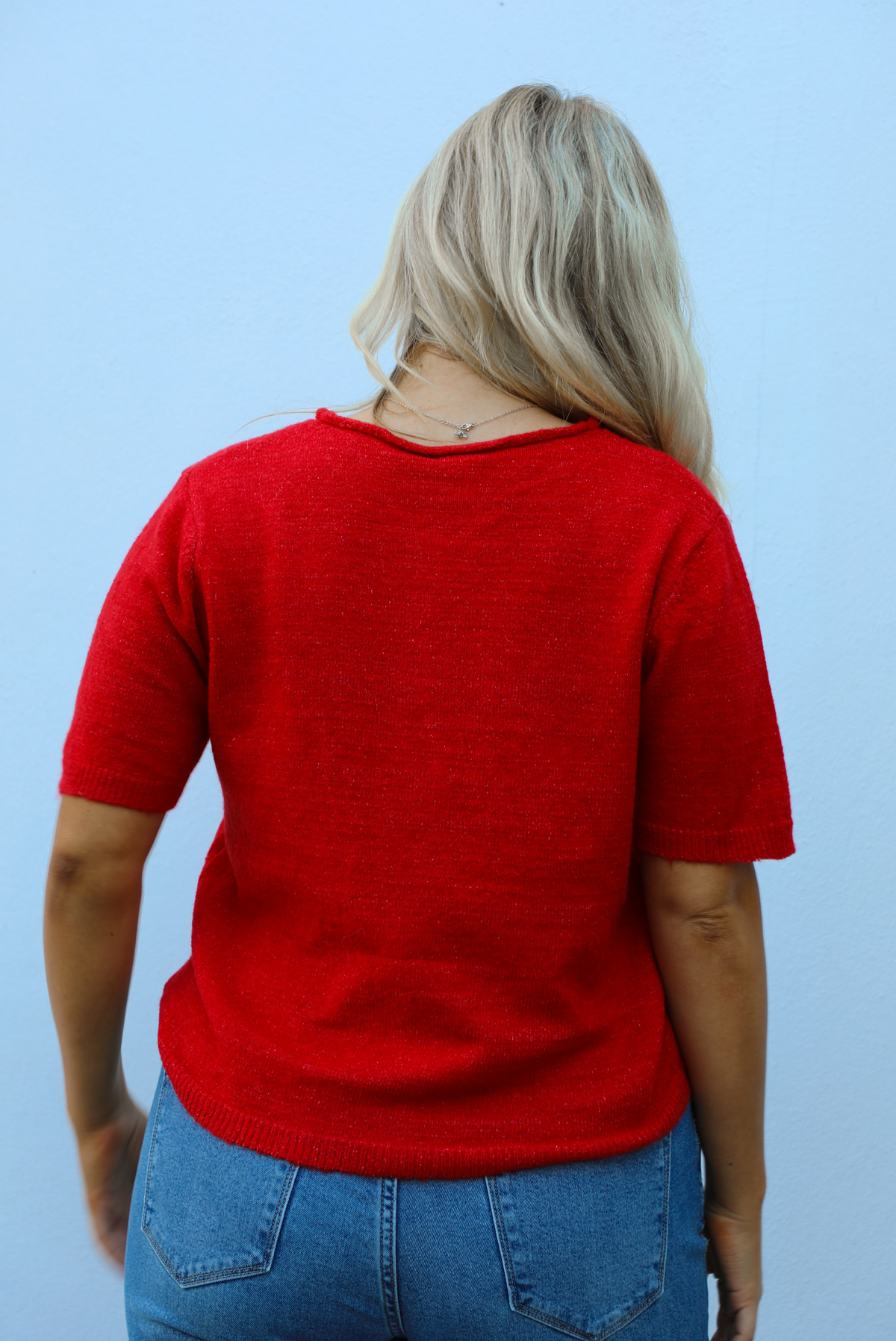 Back of a woman model wearing a red knit top that has short sleeves, a relaxed fit, and functional button closures in the front paired with a medium wash denim. The photo is taken outdoors against a white wall.