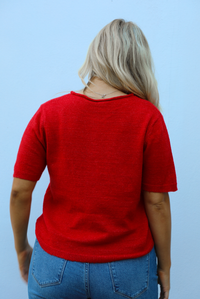 Back of a woman model wearing a red knit top that has short sleeves, a relaxed fit, and functional button closures in the front paired with a medium wash denim. The photo is taken outdoors against a white wall.