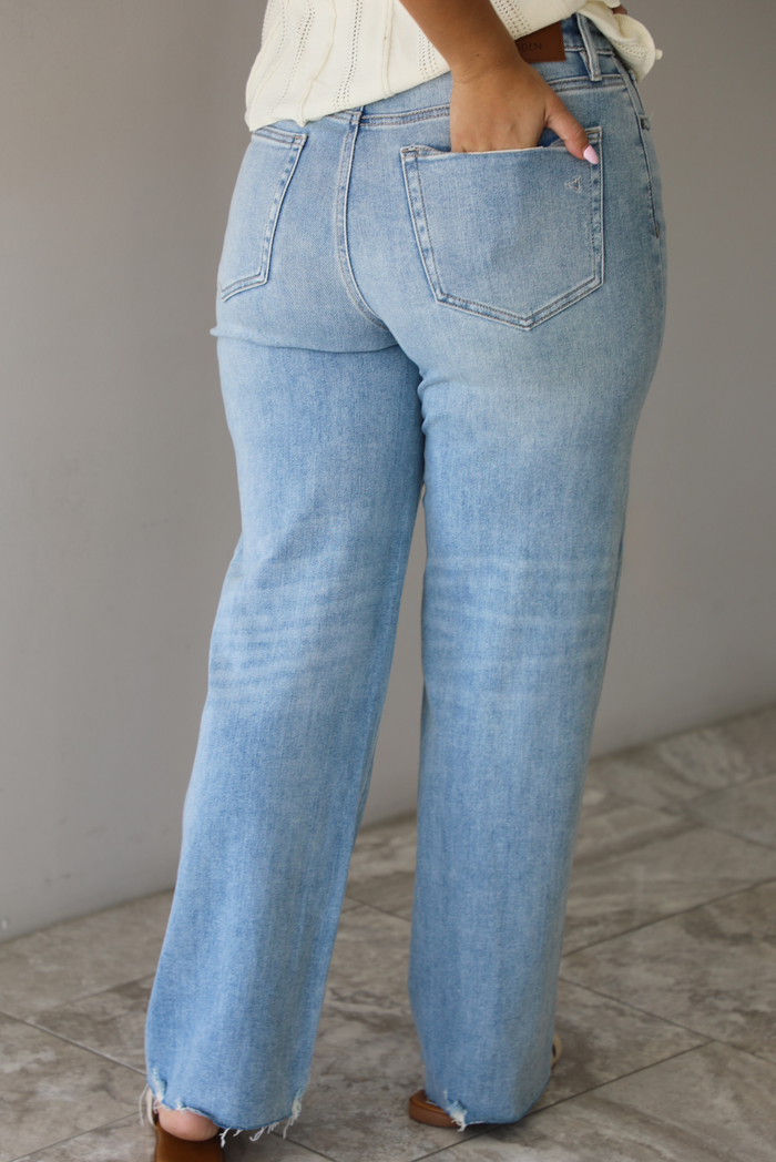 Woman wearing a pair of mid-rise light-wash denim jeans that have a raw, unfinished hem in front of a white wall with white marble flooring and shows the back of the denim. 