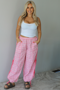 Woman wearing a white tank top with pink and white plaid pants that have a high, elastic waistline, functional pockets, and tie details on either side. The photo is taken indoors against a white wall with white marble flooring.