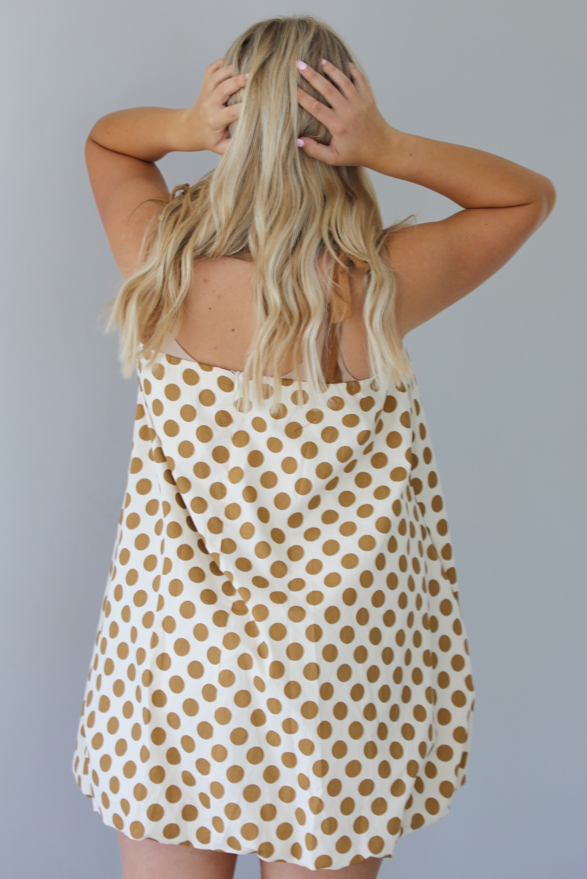 Woman wearing a cream dress with a tan polka dot print pattern throughout with adjustable tan straps, functional pockets on either side, and a bubble hem design. The photo is taken indoors against a white wall and shows the back of the dress, which has zipper closure. 