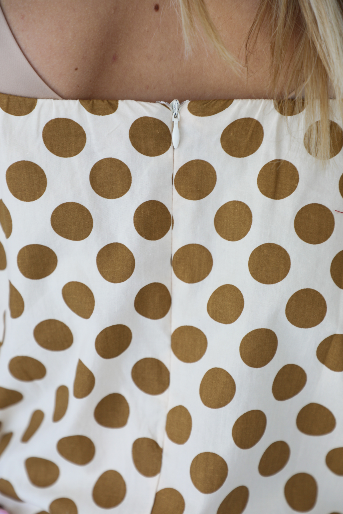 Woman wearing a cream dress with a tan polka dot print pattern throughout with adjustable tan straps, functional pockets on either side, and a bubble hem design. The photo is taken indoors against a white wall and shows a closeup of the back of the dress, which has zipper closure. 