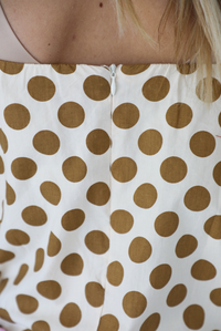 Woman wearing a cream dress with a tan polka dot print pattern throughout with adjustable tan straps, functional pockets on either side, and a bubble hem design. The photo is taken indoors against a white wall and shows a closeup of the back of the dress, which has zipper closure. 