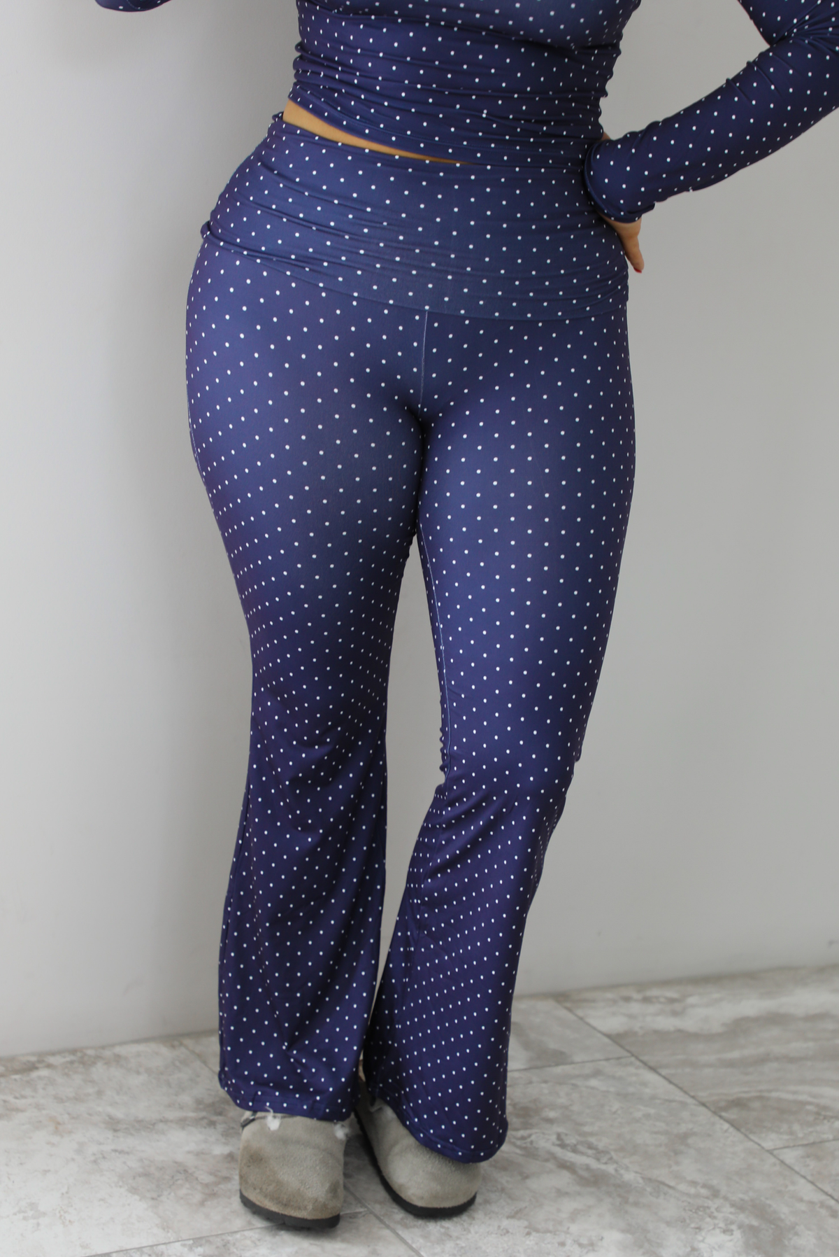 Woman wearing a navy two piece set that has a polka dot print pattern throughout. The photo shows an close up picture of the pants. The pants have a high waisted design with a fold over detail at the waistline and flared bottoms. The photo is taken indoors against a white wall with white marble flooring.