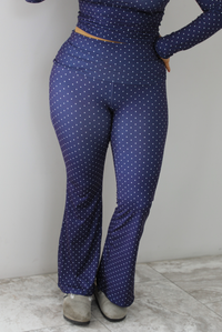 Woman wearing a navy two piece set that has a polka dot print pattern throughout. The photo shows an close up picture of the pants. The pants have a high waisted design with a fold over detail at the waistline and flared bottoms. The photo is taken indoors against a white wall with white marble flooring.