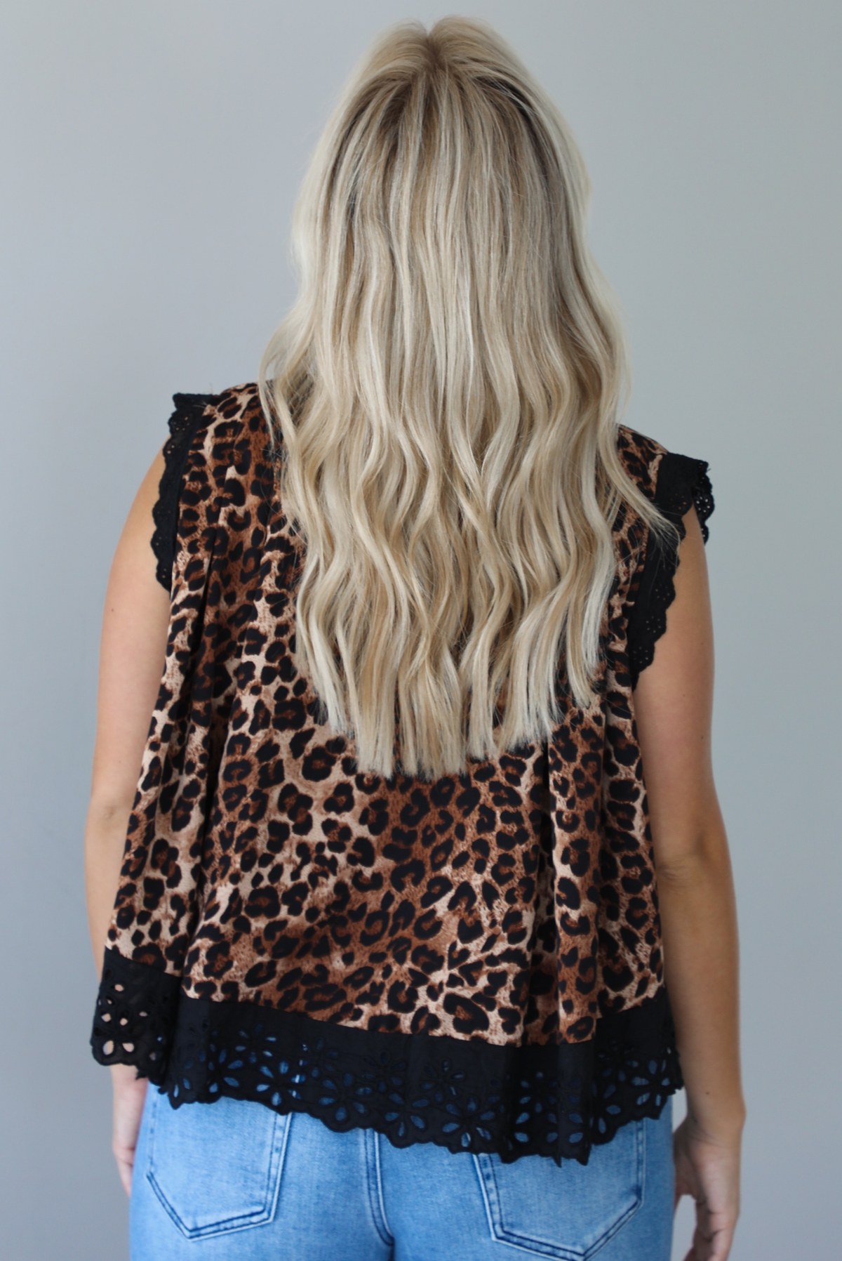 Woman wearing a black and brown leopard print flyway top featuring eyelet lace detailing at the top of the neckline, sleeves, and hem with functional button closures with a mid-rise, medium wash denim jeans indoors against a white wall and shows the back of the top. 