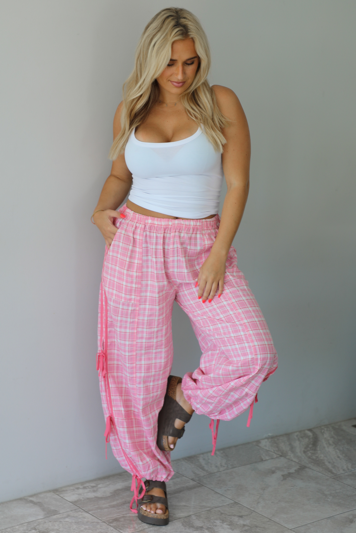 Woman wearing a white tank top with pink and white plaid pants that have a high, elastic waistline, functional pockets, and tie details on either side. The photo is taken indoors against a white wall with white marble flooring.
