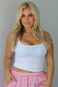 Woman wearing a white tank top with pink and white plaid pants indoors against a white wall. The photo shows a closeup of the tank, which has a ribbed texture. 