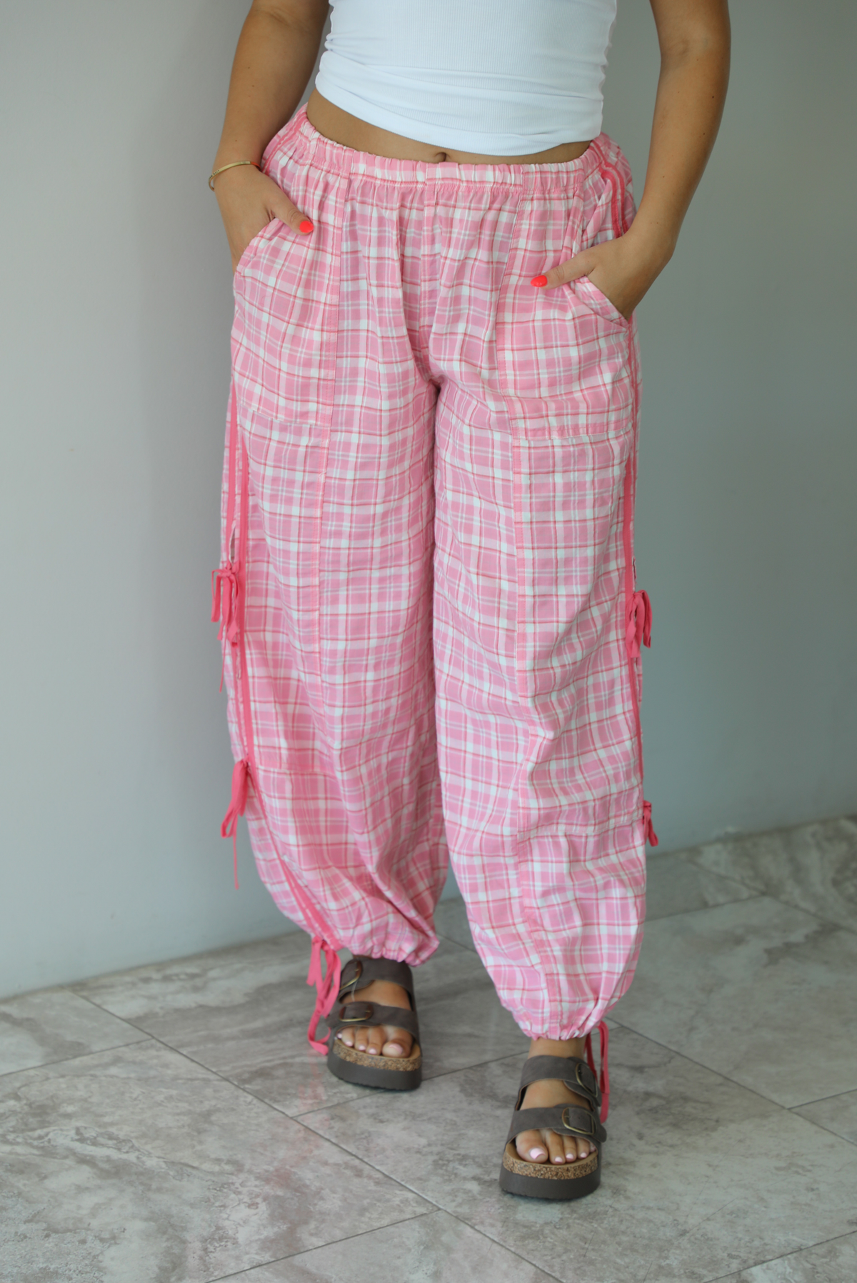 Woman wearing pink and white plaid pants that have a high, elastic waistline, functional pockets on either side, pink tie details on both sides, and are cinched with elastic at the ankles indoors against a white wall with white marble flooring. 