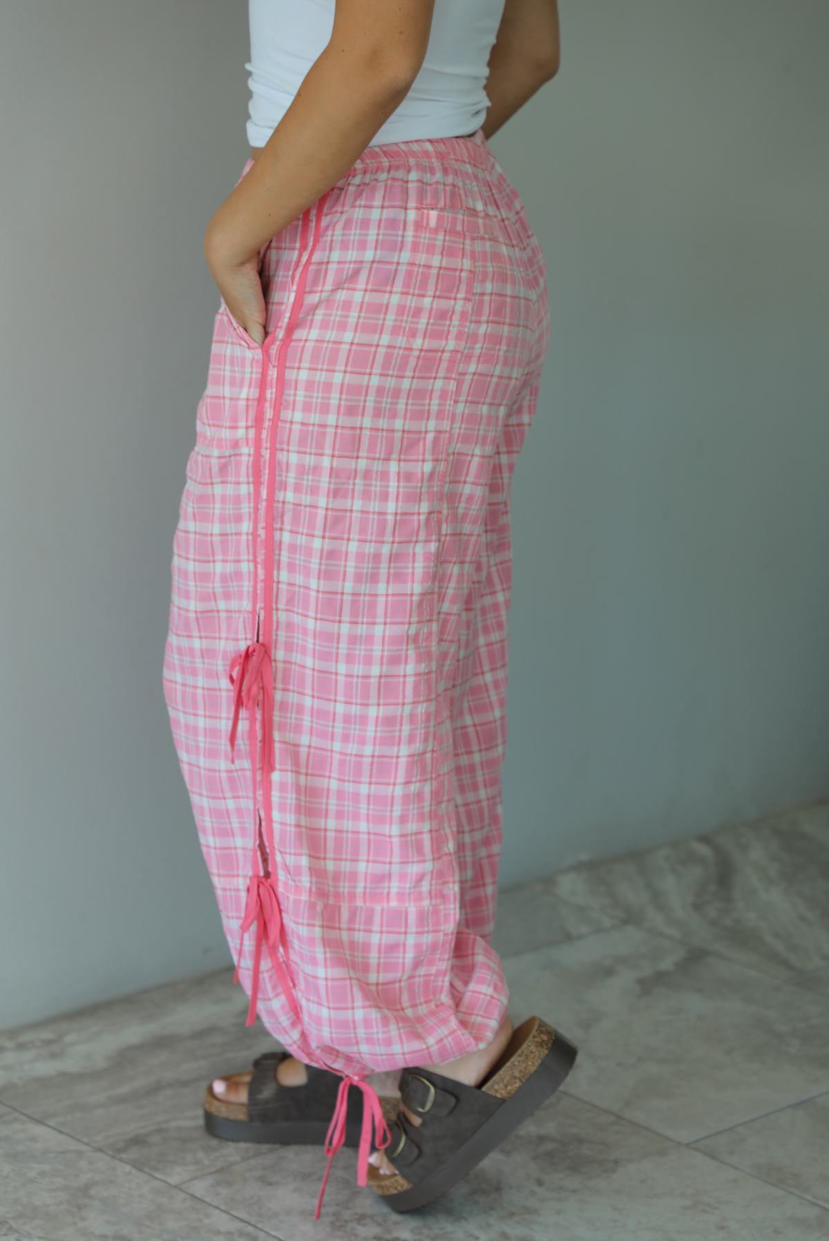 Woman wearing pink and white plaid pants that have a high, elastic waistline, functional pockets on either side, pink tie details on both sides, and are cinched with elastic at the ankles indoors against a white wall with white marble flooring. The photo shows the side of the pants, focusing on the tie details.