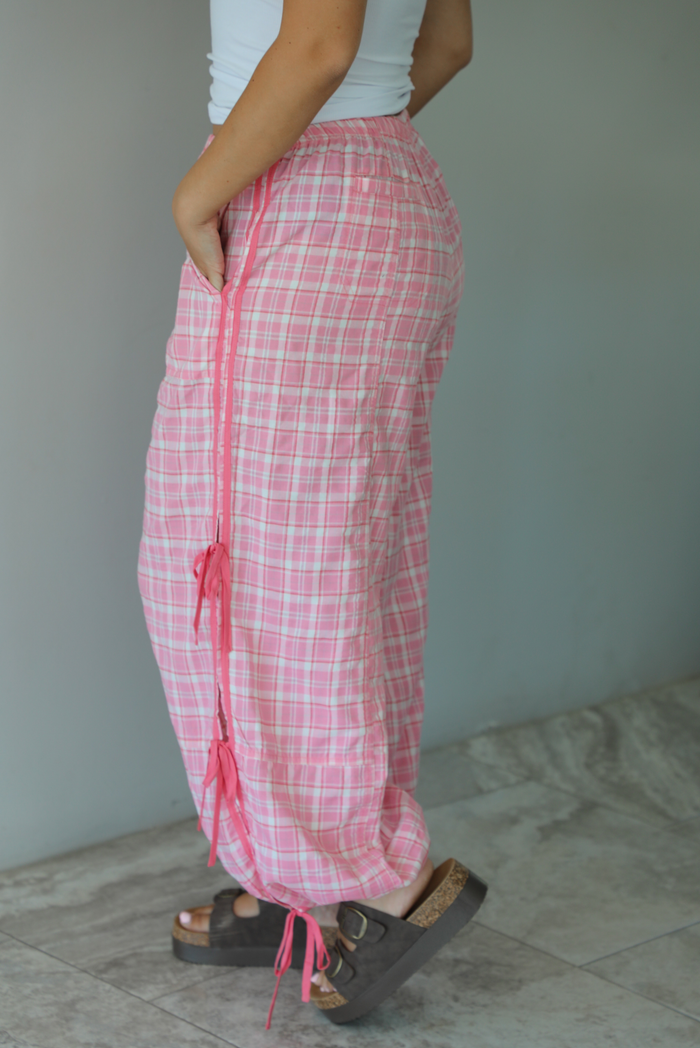Woman wearing pink and white plaid pants that have a high, elastic waistline, functional pockets on either side, pink tie details on both sides, and are cinched with elastic at the ankles indoors against a white wall with white marble flooring. The photo shows the side of the pants, focusing on the tie details.