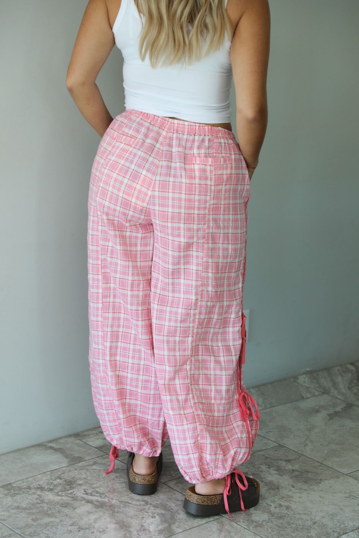 Woman wearing pink and white plaid pants that have a high, elastic waistline, functional pockets on either side, pink tie details on both sides, and are cinched with elastic at the ankles indoors against a white wall with white marble flooring. The photo shows the back of the pants. 