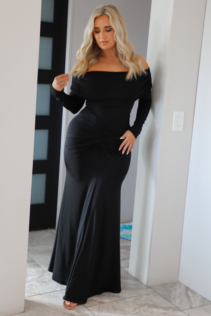 Woman wearing a fitted, black maxi dress that has a fold over detail at the shoulders, long sleeves, and a mermaid style skirt that's fitted at the hips and flows at the hem. The photo is worn indoors in a modern white home in an archway.