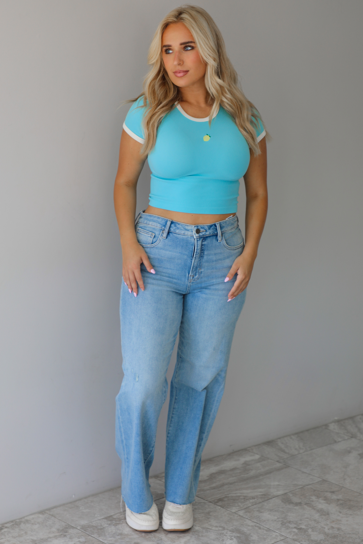 Woman wearing a blue crop top with an embroidered lemon on the front with mid-rise light-wash denim jeans against a white wall with white marble flooring. 